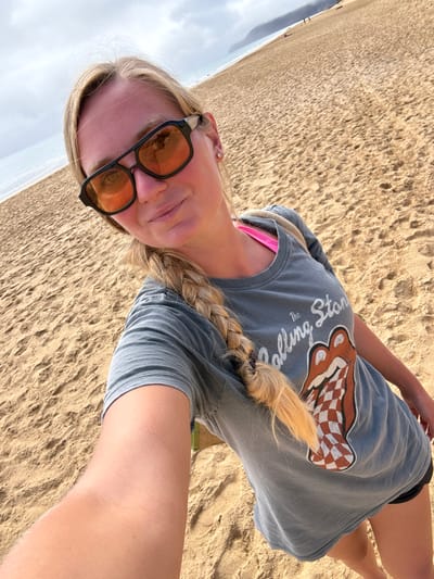 An outdoor scene on a sandy beach in Vila Baleira, Portugal, features a young adult female captured in a selfie. The female, positioned in the foreground, has light skin, long blonde hair styled in a single braid over her left shoulder, and wears black-framed sunglasses with orange-tinted lenses. She exhibits a slight smile and has a small white stud earring in her right ear. Her attire includes a faded grey short-sleeved t-shirt featuring "The Rolling Stones" text and a red and white checkered tongue and lips logo. A bright pink strap of an undergarment is visible near her neck. Portions of black shorts are visible at the bottom right. The background consists of expansive light brown sand, with the ocean to the left meeting the shoreline under a cloudy sky. In the far distance along the coastline, a prominent landmass, appearing as hills or cliffs, is discernible, with two indistinct human figures present further down the beach.