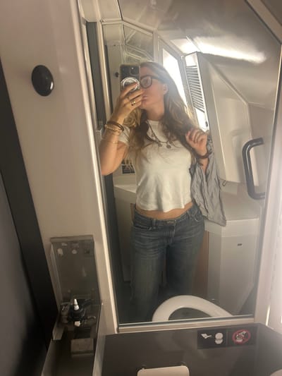 The image depicts a mirror selfie taken by a female individual within a compact, modern lavatory environment. The setting, potentially in Savigneux, France as specified, strongly suggests a public transport vehicle such as a train or aircraft due to the confined space, modular construction, and specific fixtures.
The individual, seen from the waist up, has long, wavy blonde or light brown hair and wears glasses. She is dressed in a white, short-sleeved cropped t-shirt and blue jeans, revealing a portion of her midriff. Her left arm, raised to hold a dark-cased smartphone with a circular grip, displays multiple bracelets (some wooden, some metallic) and a ring on her ring finger. Her lips are puckered in a "kiss" pose. Her right hand is seen holding a light-colored, possibly striped, piece of fabric over her right shoulder, with fingers also touching her hair.
The mirror reflects the individual and the surrounding lavatory. To the left of the reflection, a dark panel features a circular black button or knob, below which is a metallic recess containing a dispenser (likely soap or sanitizer) and other fixtures. A white toilet seat is visible in the lower center of the reflection. Below the toilet seat, two international pictograms are present: one depicting a person sitting on a toilet, and another with a crossed-out figure standing on a toilet, indicating a prohibition. To the right of the reflection, a black grab bar is mounted on a white wall. The overall environment is brightly lit and features white and grey modular surfaces.