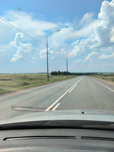 The image depicts a straight, paved road with white lane markings, viewed from the perspective of a vehicle's dashboard. The road stretches towards a distant horizon lined with trees. On either side of the road are grassy fields with scattered utility poles. The sky is blue with prominent, fluffy white clouds. Two tall lamp posts are visible along the right side of the road. The foreground shows the hood of a car, suggesting the image was taken while driving. The overall scene suggests a rural or semi-rural setting. While specific landmarks for Старокутлумбетьево, Russia, are not identifiable, the environment is consistent with the general landscape of such a location.
