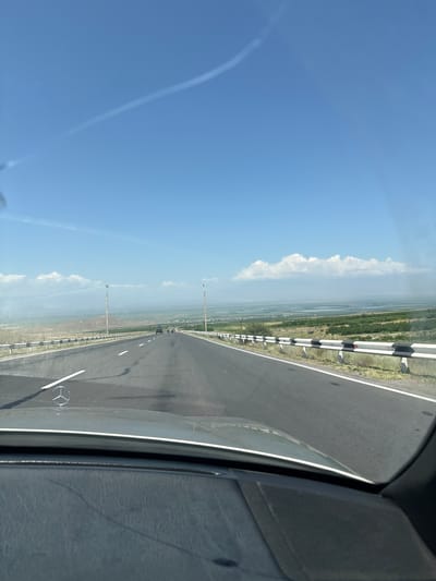 The image captures a view from inside a vehicle, looking forward along a highway. The road is asphalt, with white lane markings. A metal guardrail with black and white reflective markers lines the right side of the highway. The landscape on either side of the road is flat and appears to be agricultural, with some distant hills and bodies of water. The sky is a clear blue with some wispy white clouds. Two cars are visible on the highway in the distance. The hood of the vehicle in the foreground is visible, with a Mercedes-Benz emblem. The scene is set in the city of Paruyr Sevak, Armenia.