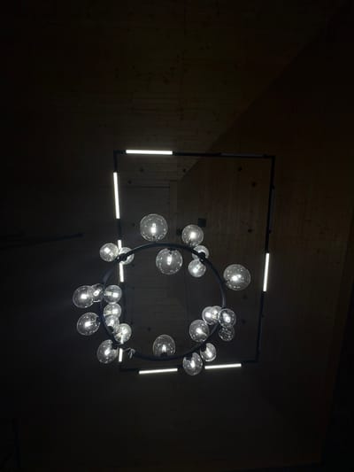 The image depicts a modern chandelier hanging in a dimly lit space. The chandelier has a circular, black metal frame with multiple illuminated glass globes attached around its circumference. A rectangular black metal frame surrounds the circular element, and integrated into this frame are four linear LED light bars. The walls appear to be wood paneled, suggesting an interior space. The low lighting levels and absence of other identifying features make it difficult to determine the exact location, but the prompt indicates it is in Kamennoye, Russia. There are no people visible in the image.