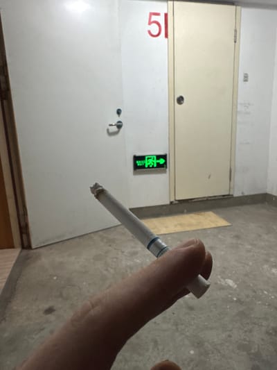 A partially lit cigarette is held by a person's hand in the foreground. The cigarette is angled upwards and to the left, with a small ember at the tip. In the background, there are two doors, one white and one beige, with a red number "51" painted on the wall above the beige door. A green "EXIT" sign with an arrow pointing to the right is mounted on the wall between the two doors. The floor is made of concrete, and the environment appears to be a hallway or corridor, possibly in a residential building or parking garage. The lighting is dim.