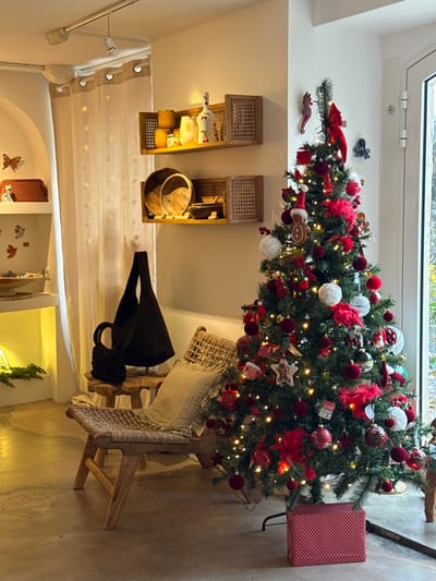 The image depicts an indoor commercial space, likely a boutique or gallery, located in Saint-Paul-de-Vence, France, decorated for the Christmas season. The environment features white walls and a light-colored floor, possibly concrete.

To the right, a tall, artificial Christmas tree stands, illuminated by numerous warm white string lights. It is heavily adorned with predominantly red and white ornaments, including various baubles, fluffy pom-poms, red feather accents, star-shaped ornaments, and a gingerbread man figure wearing a Santa hat. A red seahorse ornament and a red ribbon bow are visible near the tree's apex. At the base of the tree, a single, rectangular gift box wrapped in red and white polka-dotted paper is present. A large glass window or door on the far right provides ambient natural light.

In the center-left foreground, a rustic, light-toned wooden lounge chair with a woven seat and back is positioned, holding a striped decorative cushion. Adjacent to the chair, a small, irregular wooden stool with three legs supports two black bags: a large, soft tote bag with long handles draped over it, and a smaller, woven or crocheted handbag with two top handles.

Above the wooden stool, two rectangular floating wooden shelves with integrated woven cane panels are mounted on the wall. The upper shelf displays decorative items including yellowish candle-like objects, a white vase with red cherry motifs, and other small artifacts. The lower shelf holds several wooden bowls and other natural material items.

Further to the left, a white arched alcove serves as a display area, featuring tiered white shelves. These shelves showcase various craft items, including a woven red clutch, wooden butterfly cutouts, ceramic bird figures, and a glowing light source nestled within a green garland on the bottom shelf.

The ceiling above features a white track lighting system with at least one visible spotlight directed towards the displays. A light-colored, sheer curtain with small circular patterns hangs on the far left. No people are present in the scene.