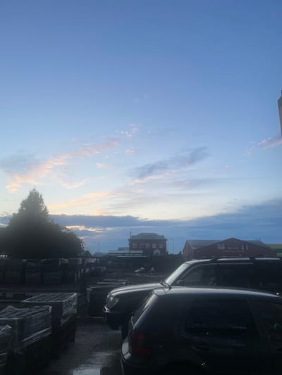 The image depicts a street scene in Oral, Kazakhstan, likely during the early morning or late evening, given the twilight sky. In the foreground, two cars are parked on a wet paved surface. The car on the right is a dark-colored hatchback, while the car to its left is a larger, dark-colored SUV. Behind the vehicles, stacks of building materials, possibly bricks or paving stones, are arranged on pallets and wrapped in plastic. A large evergreen tree stands to the left, silhouetted against the sky. In the background, several buildings are visible, including a brick building with multiple windows and a larger, red-roofed structure with signage that appears to be in a Cyrillic alphabet. The sky above is a gradient of light blue with scattered clouds tinged with pink and orange, suggesting the sun is either rising or setting. The overall impression is one of an industrial or commercial area at the edge of the city, with a focus on construction materials and vehicles.