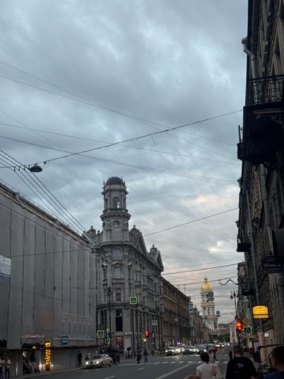This image captures a view of a street in Saint Petersburg, Russia, likely near the Kryukov Canal or on the approach to St. Nicholas Naval Cathedral. The sky is overcast with dramatic, grey clouds, suggesting either early morning, late evening, or impending rain.
In the foreground, a light-colored sedan is driving on the wet asphalt road. People are visible on the sidewalk to the left, some standing and some seated, under a makeshift shelter with illuminated lights. A building on the left is covered in a large, grey tarp, possibly undergoing renovation.
Further down the street, an ornate, stone building with a distinctive tower and dome is a prominent feature. Across the street, the façade of a classical building is visible. Several traffic signals are present, including a red traffic light. Signs with Cyrillic script are mounted on poles and building facades. The street is lined with apartment buildings, some with balconies. Overhead, a complex network of wires crisscrosses the sky, a common sight in many older urban areas.
In the distance, the golden dome of a church, likely St. Nicholas Naval Cathedral, gleams through the cloudy sky, serving as a landmark. The overall atmosphere is urban and somewhat muted due to the weather conditions, yet the architectural details of the buildings and the distant golden dome suggest the historical and cultural richness of the city.