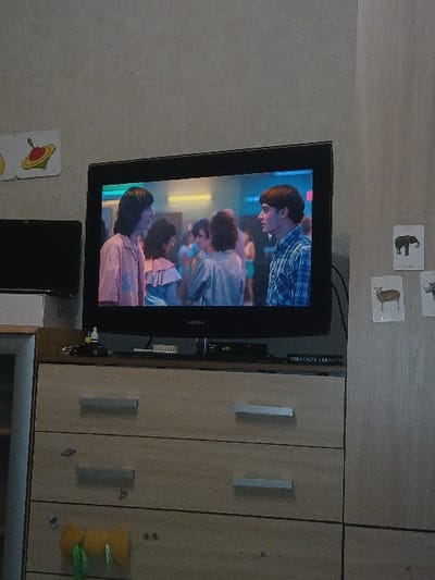 A television displaying a scene with multiple people is mounted on a wall beside a wooden cabinet with silver handles. Playing cards with animal illustrations are placed on the wall near the TV. A black box, likely a DVD player, is positioned underneath the TV. More playing cards are near the bottom of the screen, and a stuffed animal hangs off the side of the cabinet.
