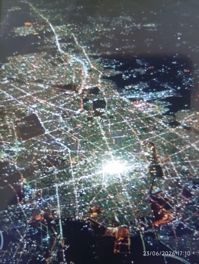 This media file captures an aerial view of a city at night, likely Eshanguzar, Uzbekistan. The scene is dominated by the intricate network of city lights, forming geometric patterns across the landscape. Roads or pathways are illuminated by the lights, crisscrossing the urban sprawl. Buildings are represented by clusters of lights, some appearing denser and brighter, suggesting more developed areas or prominent structures. The overall impression is one of a vibrant, active city at night.
The time of day is clearly night, indicated by the darkness of the sky and the prominence of artificial lighting. There are no visible cues about the weather, but the clear visibility of the lights suggests it is not heavily overcast or foggy. The lighting itself provides the primary visual interest, with a particularly bright, almost blinding light source in the central-lower portion of the image, which could be a stadium, a large industrial complex, or a powerful searchlight.
Some darker, less illuminated areas are also visible, potentially parks, undeveloped land, or bodies of water. The texture of the image suggests it might be a photograph taken from a considerable height, possibly from an airplane or a drone, given the wide expanse of the city visible.
The visible text at the bottom right corner of the image reads "23/06/2026 17:10". This timestamp, given the visual context of nighttime, is curious. It's possible this is a future date, or the timestamp is inaccurate or belongs to a different event. Regardless, it provides a specific date and time reference associated with the media. There are no people or specific objects readily identifiable beyond the urban infrastructure itself. The scene evokes a sense of scale and the human endeavor of building and illuminating a city.