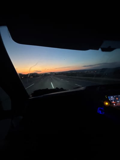 A car is driving on a highway at sunset. The sky is a gradient of orange, pink, and blue. The highway has multiple lanes and is lined with streetlights. There are some hills in the distance. The interior of the car is dark, but the dashboard lights are visible. The image is taken from the perspective of the driver.