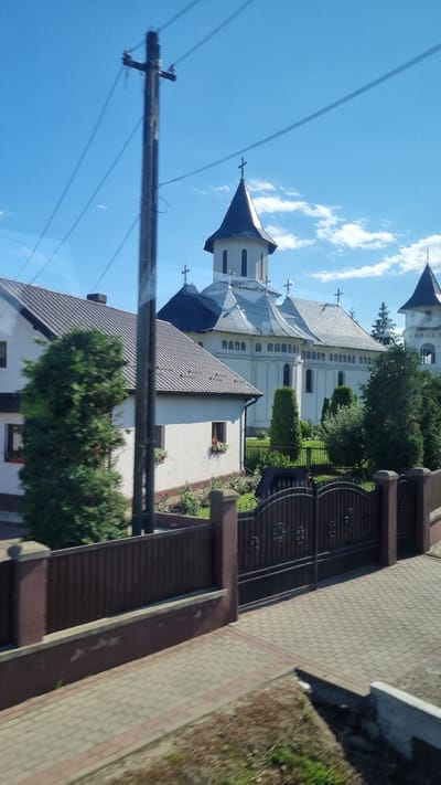 The image depicts a large, white, multi-turreted church with multiple crosses, characteristic of Orthodox Christian architecture, situated in Milișăuți, Romania. The church features a prominent central tower with a dark, conical roof topped by a cross. To the left of the church, a smaller white building with a grey tiled roof is visible, partially obscured by a tall, dark wooden utility pole with wires running across the frame. A dark metal gate with decorative elements stands in front of the church grounds, with a brick pathway leading away from it. The environment appears to be a clear, sunny day with a blue sky and scattered white clouds. The overall scene conveys a sense of tranquility and religious solemnity.