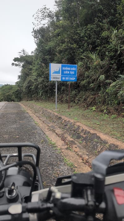 The image shows a road sign in Thượng Trạch Commune, Vietnam. The sign is blue and white and displays Vietnamese text and a graphic of a downward sloping road. The text on the sign reads "XUỐNG DỐC LIÊN TỤC" which translates to "Continuous Descent" and "13950m ĐI SỐ THẬP" which translates to "13950m Go to Low Gear". The sign is placed on the side of a paved road that curves to the left and is surrounded by lush green vegetation and trees. In the foreground, part of a motorcycle is visible, suggesting the photo was taken by a rider. The sky is overcast.