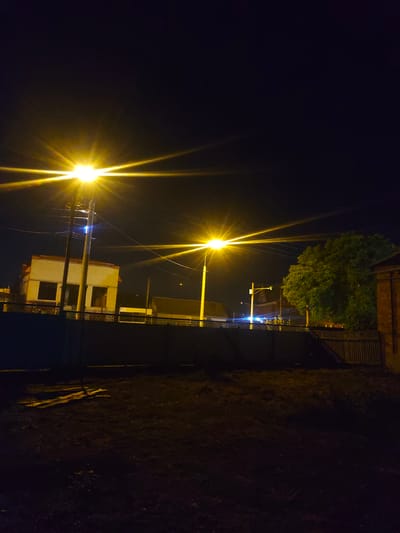 The image depicts a nighttime urban scene, possibly in Novoukrainskoe, Russia, illuminated by streetlights. A bright, starburst-like streetlight dominates the left side of the frame, casting a yellowish glow. In the middle ground, a building with a yellow facade and dark windows is visible, with electrical poles and wires in front of it. To the right, a large, leafy green tree stands, also lit by the streetlights. A dark fence runs across the middle ground, separating the illuminated area from a darker foreground which appears to be a patch of ground with some debris or discarded materials. The sky is dark and clear, with no visible moon or stars apart from what appears to be a distant speck in the upper right.