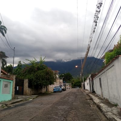 The image shows a residential street in El Limón, Venezuela, under a cloudy sky. A narrow, paved road with cracked asphalt runs uphill, lined with houses and stone walls on either side. On the left, a light blue car is parked in front of a turquoise house with a white-barred window. Further up the street, a white SUV and another vehicle are parked. A large, bushy tree with pink flowers dominates the left side of the frame. On the right, a white concrete wall with horizontal lines runs along the street, with a doorway visible. Overhead, numerous electrical wires crisscross the sky, some of them adorned with epiphytic plants. In the background, a mountain range is shrouded in dark, low-hanging clouds. A street lamp with an orange glow is visible on the right side of the road.