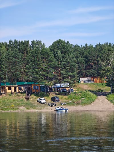The image depicts a serene waterscape with a forested background and a small settlement along the bank. A wide, calm river occupies the foreground, its surface rippling and reflecting the green of the trees and the blue of the sky. The far bank is lined with a dense forest of tall pine trees.
Nestled at the base of the forest, on a slightly elevated grassy embankment, are several wooden structures, likely small houses or cabins, with green roofs. In front of these structures, a sandy area slopes down to the water. Several vehicles are parked on the embankment, including a white SUV towing a boat trailer and a black SUV. A small motorboat is docked near the shore. A wooden staircase leads up from the sandy area to one of the buildings. The sky above is a clear, bright blue with a few wispy clouds. The overall impression is one of a peaceful, rural setting, possibly a recreational area or a small village on the outskirts of Podporozh'e, Russia, given the context of the query.