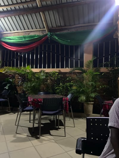 The image depicts a nighttime scene in an outdoor or semi-outdoor dining and relaxation area, likely in Bagamoyo, Tanzania.
The setting features a rustic ceiling composed of wooden beams supporting corrugated metal sheets. From this structure, green and red fabric drapes are tied around a wooden support, adding a decorative element. A bright light source emanates from the upper left, casting a strong glare and illuminating parts of the ceiling and drapes.
In the midground, a light-colored wall or partition acts as a backdrop for several lush green potted plants, including large palm-like foliage, contributing to a tropical ambiance. Behind this wall, a darker area with horizontal light lines suggests a fence or railings.
The foreground showcases a seating arrangement with multiple tables and chairs. The tables have dark surfaces, possibly wood or glass, supported by metallic frames. The chairs feature dark, perforated backs and seats, also with metallic frames. Some chairs are adorned with red and black plaid fabric, likely decorative throws or seat covers. A single person is partially visible in the upper right, seated at one of the tables, wearing a light-colored, patterned shirt.
The ground is paved with light-colored square tiles, which reflect the ambient light and clearly display the distinct shadows cast by the tables and chairs, indicating strong illumination from overhead or the side. The overall setup suggests a casual dining or social gathering space. There is no visible text in the image.