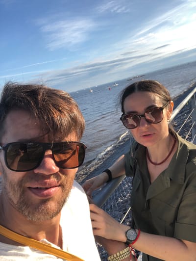 A selfie is taken by a man and a woman who are standing next to each other on an embankment. The man is in the foreground, wearing sunglasses, and the woman is behind him, also wearing sunglasses. They are both looking at the camera. The background shows a body of water with several sailboats on it, and a blue sky with clouds. The location is identified as Zmeinyi, Russia. The man has brown hair and a beard, and he is wearing a white shirt. The woman has dark hair and is wearing a green shirt and a red necklace. There is a railing in front of them. The image is taken in daylight.