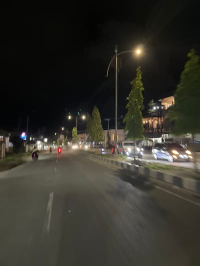 The image captures a night scene on a street, possibly in or near the city of Dalam, Indonesia. The perspective is from a moving vehicle, resulting in motion blur throughout the frame. On the right side of the road, there are several parked cars, one light-colored SUV and two darker sedans, their headlights illuminated. The street itself is asphalt with painted lane markings and a concrete divider with alternating black and white reflective blocks.

To the left of the road, buildings are visible, some with illuminated windows and signage. Several trees, appearing to be tall and coniferous, are planted along the roadside and in front of the buildings. A streetlamp with a bright yellow light is positioned in the upper middle of the frame, casting a glow on the surroundings.

Further down the road, other vehicles, including motorcycles and cars, are in motion, their lights creating streaks of color due to the motion blur. One motorcycle on the left side of the road is particularly visible, with its rider illuminated by the headlights. The sky above is dark, indicating nighttime. The overall impression is of a moderately lit urban or suburban street at night.