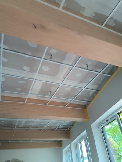 The image shows a room under construction. The ceiling has a grid of white metal supports, with drywall partially installed and patched. Wooden beams run across the top and bottom of the view. One beam has a visible knot. Windows are present on the right side of the image, with trees visible outside. The walls are white. Yellow tape is visible along the edge of the ceiling. Based on contextual clues and architectural style, the location is likely Münster, Germany.