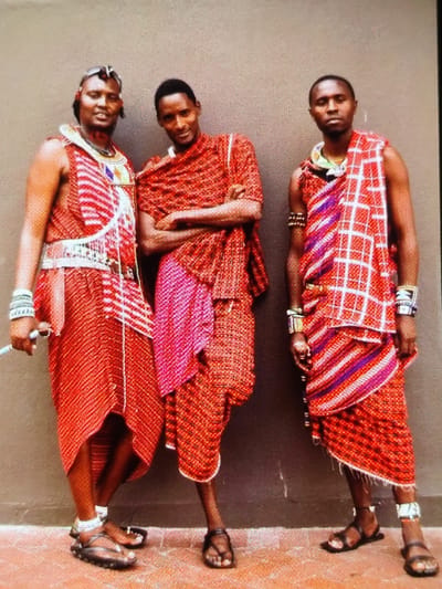 Three adult males stand in a frontal pose against a neutral-toned wall on a textured, reddish-brown ground in Ihumwa, Tanzania. All individuals wear traditional garments, primarily red patterned fabrics, along with various beaded ornaments and dark sandals.
The leftmost male, facing forward, displays a faint smile. He wears a prominent white beaded chest ornament, a white beaded belt, numerous white wrist bracelets, white beaded anklets, and dark sandals. He holds a slender, light-colored object in his left hand.
The central male, facing forward with a faint smile and arms crossed over his chest, is draped in red patterned fabric over a visible pink inner layer, and wears dark sandals.
The rightmost male, facing forward with a neutral expression, wears a multicolored beaded necklace, multiple beaded bracelets on his left wrist, silver-toned bracelets on his right wrist, white beaded anklets, and dark sandals.