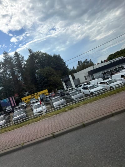 The image shows an outdoor car dealership in Wieliczka, Poland. A variety of cars, including sedans, SUVs, and vans, are parked in rows behind a fence. Some of the cars are white, while others are darker colors. A yellow sign with Polish text, "NAPRAWY BLACHARSKO-LAKIERNICZE" (Bodywork and paint repairs), is visible. To the right, a building with "Renault SERWIS" signage indicates a Renault service center. The sky is cloudy with patches of blue. Trees surround the dealership. A paved sidewalk and road are in the foreground.