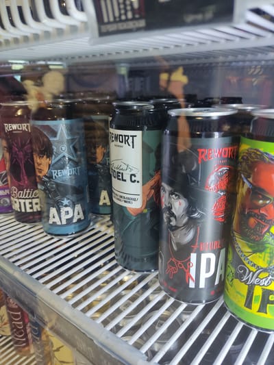 The image shows a shelf in a refrigerator stocked with cans of beer from Rewort Brewery. The beers include Rewort Baltic Porter, Rewort APA, Rewort Del C., Rewort Double IPA, and West IPA. The location is likely in a shop or establishment in Pskov, Russia, where Rewort Brewery is located. The refrigerator has white wire shelving.