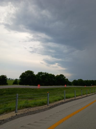 The image shows a view of a highway on a cloudy day. The road has a yellow line marking the edge. To the left of the road is a grassy median with orange traffic cones and a wire fence. Trees line the median. The sky is filled with gray clouds. The image appears to have been taken during the daytime, possibly late afternoon or early evening given the cloud cover. There is no visible text in the image. The scene suggests a rural or semi-rural location, consistent with being in or near Lexington, United States. There are no people or specific activities depicted.