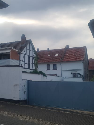 The image shows a partial view of a building facade with several architectural styles. The main structure is white with a tiled red roof and some timber framing on a gabled section. There are several windows visible, some with dark frames and others with lighter frames. A section of the building is covered by a large, dark blue, flat surface, possibly a modern addition or a facade. To the right of this blue surface, a brick wall is visible, and further right, a paved area that appears to be a sidewalk or street. The sky is overcast with a bright streak of light, suggesting either a sunset, sunrise, or a break in the clouds. The scene appears to be in daylight, likely late afternoon or early morning given the light. There is some greenery, possibly ivy, growing on the white wall. The overall impression is of an urban or semi-urban environment with a mix of old and new architecture. No people are visible in the frame. The orientation of the image is rotated, with the sky appearing on the left and the ground on the right.