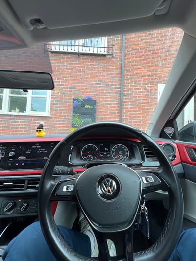 From the driver's seat of a Volkswagen, a brick building with a window box filled with purple and green plants is visible. A yellow rubber duck wearing a black beanie sits on the dashboard. The car's digital display shows "11:48" and various statistics. A side mirror and sun visor are visible in the foreground.