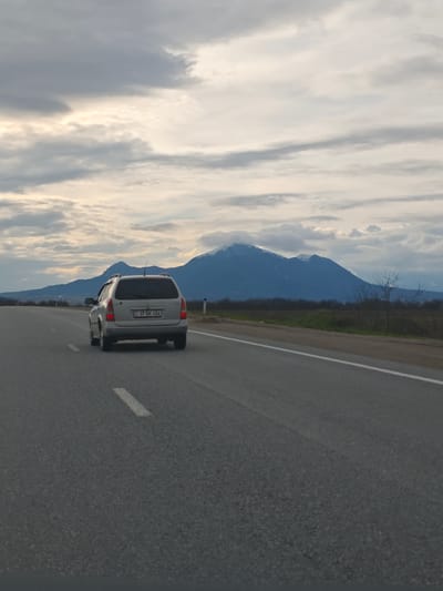 A silver station wagon travels on an asphalt road with a dashed white line. Mountains with snow-covered peaks are visible in the background under a cloudy sky. The car's license plate reads "37 BK 456." The road is flanked by vegetation on the side. The location is Konstantinovskaya, Russia.