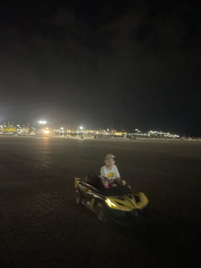 In Merchanskoe, Russia, a young child wearing a baseball cap and a white t-shirt with a yellow design is seated in a yellow toy car. The car is on a paved surface that appears to be cobblestone. In the background, a blurred panorama of city lights and structures suggests a waterfront or harbor area. The scene is captured at night, with the sky appearing dark and the lights in the distance creating streaks and blurs due to motion or low light conditions.