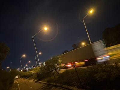 The image depicts a night scene along a highway in Bull Creek, Australia. A large semi-trailer truck is captured in motion, its headlights and taillights creating streaks of light due to motion blur. The truck is traveling on a multi-lane highway, indicated by the presence of streetlights and an "EXIT" sign with an arrow pointing right. The surrounding environment includes a chain-link fence, dense vegetation, and a pathway, possibly a bike or pedestrian lane, illuminated by streetlights. The sky is dark, characteristic of nighttime, with some streetlights casting a yellow glow and creating lens flares in the camera. The overall impression is a dynamic highway environment at night.