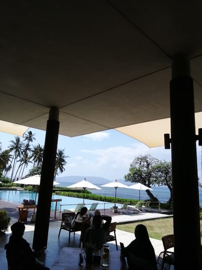 The image depicts a tropical resort scene in Banyuwangi, Indonesia, featuring an outdoor seating area with patrons enjoying a view of the ocean and a swimming pool. The perspective is from under a covered patio, with large support pillars and a ceiling structure dominating the foreground.
In the foreground, silhouetted figures are seated at tables. On the left, a person is seated facing left, looking down. In the center, another person is seated at a table, looking at a phone. To the right, a person wearing a hijab is seated at a table, also looking at their phone.
Beyond the seating area, a large swimming pool is visible, lined with lounge chairs and umbrellas. Palm trees line the background, with a lush green landscape leading to the ocean. The ocean itself is calm, with distant islands or landmasses visible on the horizon under a bright, partly cloudy sky. White umbrellas are strategically placed around the pool area, offering shade. The overall impression is one of a relaxed, upscale resort atmosphere.