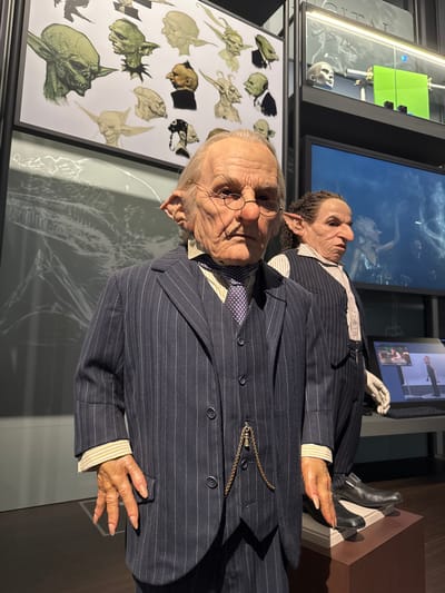 This photograph depicts a display of animatronic figures in what appears to be a museum or exhibition. The subjects are likely goblins or similar fantastical creatures, dressed in pinstripe suits with pocket watches, suggesting an Edwardian or early 20th-century aesthetic. The figures have detailed facial features, including one with glasses, which suggest careful craftsmanship and attention to realism.

The background features conceptual sketches of similar creatures on a framed poster and behind that a translucent drawing on a glass panel, adding context and showing the design process. In the upper portion of the image, several screens are visible.

The exact location cannot be determined to be Nerima, Japan, based solely on the visual information provided.