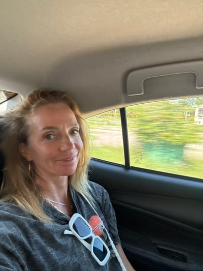 An adult female is depicted seated inside a moving passenger vehicle, likely in the back seat, within the city of Panglao, Philippines. Her blonde hair is loosely styled, and she is looking directly at the camera with a slight smile. She is wearing a dark grey t-shirt featuring a graphic design of a red circle with a white outline. A silver chain necklace and hoop earrings are visible. A pair of white-framed, rectangular sunglasses with dark lenses hangs on a white strap around her neck, resting on her chest. The vehicle's interior features a light-colored headliner and a grey grab handle above the window. Through the side window, a motion-blurred green landscape with some light-colored structures indicates the vehicle is in transit.