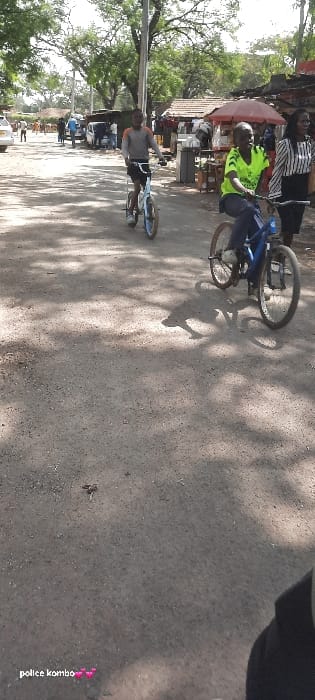 The image depicts an outdoor daytime scene on an unpaved road in Homa Bay, Kenya. Two individuals are actively riding bicycles in the mid-ground. The subject on the left, wearing a grey long-sleeved shirt and dark shorts, operates a light blue bicycle. To their right, a second subject, in a bright green short-sleeved shirt and dark trousers, rides a blue bicycle equipped with a water bottle cage on its frame. Adjacent to this second cyclist and slightly behind, a female subject wearing a black and white striped long-sleeved shirt and dark lower garment walks to the right.
The road surface consists of dirt or gravel, showing varied texture and small scattered debris. Numerous dappled shadows from overhead trees are cast across the road, indicating direct sunlight from above and to the right. In the background, along the right side of the road, a series of market stalls or small, rudimentary shops are present, featuring corrugated iron roofs and at least one prominent red umbrella acting as an awning. A tall, dark utility pole stands vertically in the center-left mid-ground. Further in the background, on the left side, a light-colored sedan is visible, along with several indistinct individuals walking or standing. The immediate foreground shows a small portion of a dark garment on the bottom right.
