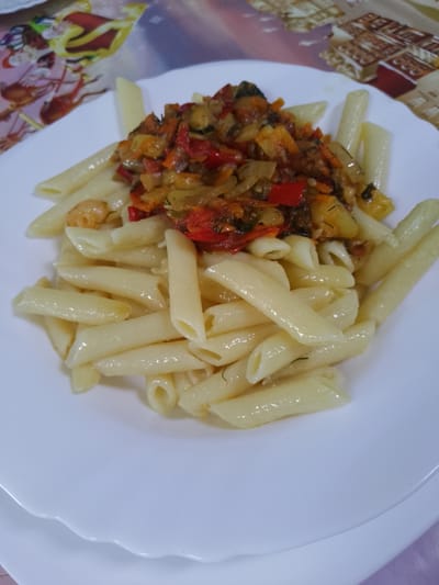 A close-up, top-down view of a white, scalloped plate filled with penne pasta and a topping of sautéed vegetables. The pasta is cooked al dente and appears to be coated in a light oil. The vegetable topping is a colorful mix of diced red bell peppers, zucchini, carrots, and onions, with visible flecks of dill. The background shows a patterned tablecloth with a vintage aesthetic, possibly a placemat or tablecloth, in shades of beige and red with an illustration of what looks like old books. The lighting is natural and suggests an indoor setting. There are no people visible in the image. The scene depicts a prepared meal. The location is not identifiable in the image itself.