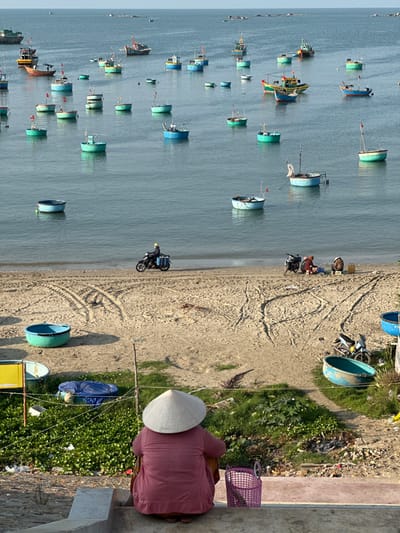 A beach landscape features numerous colorful fishing boats in the calm, blue water. A sandy beach runs alongside, showing tire tracks and parked motorcycles. At the edge of the beach, a person wearing a conical hat and a pink outfit stands near a small building. Green vegetation and scattered debris are visible nearby. The overall scene presents a blend of maritime activity and coastal life.