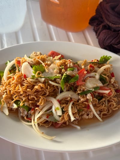 A partially filled white plate of Yum Mama, a Thai noodle salad, sits on a white table. The dish contains instant noodles, sliced onions, tomatoes, green vegetables, and ground pork. An orange container and a brown cloth are to the left of the plate. The photo was taken in Na Kluea, Thailand.