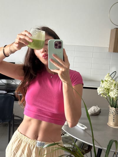 A young woman with dark hair, wearing a bright pink crop top and striped yellow pajama bottoms, is taking a mirror selfie in what appears to be a kitchen. She is holding a clear glass filled with a green smoothie in her left hand and a light green smartphone in her right hand, which is angled to capture the reflection. Her lips are pursed as if she's about to take a sip. In the background, a vase with white flowers sits on a table next to a remote control and other kitchen items. The environment suggests a domestic setting, likely in Tel-Aviv, Israel, given the context.