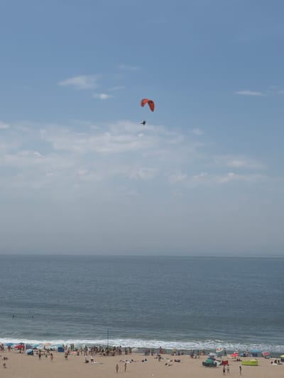 The image shows a sunny day at a crowded beach in Espinho, Portugal. A person is paragliding in the bright blue sky, with the vast ocean to the right. The beach is filled with people sunbathing, swimming, and relaxing under colorful umbrellas. The waves are crashing gently on the shore, creating a peaceful atmosphere. The time of day appears to be late morning or early afternoon, as indicated by the bright sunlight and long shadows. The weather is warm and pleasant, perfect for a day at the beach.