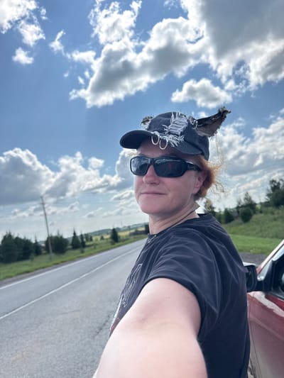 An outdoor, medium shot shows a person standing next to a red vehicle on a rural road. The person is wearing a black t-shirt, sunglasses, and a distressed denim baseball cap adorned with shredded fabric and metal rings. A small bird is perched on the cap. The person has reddish-brown hair, and their face is partially obscured by the sunglasses. They are holding a phone to take a selfie, with their arm extended. The road stretches into the distance, bordered by green trees and fields. The sky is a bright blue with scattered white clouds. The scene is captured during daylight. There is no indication of the location being in the city of Рождественск, Russia.