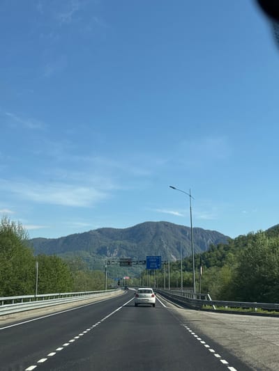 The image depicts a road in Monastyr', Russia. A white car travels on the right lane of the road. A mountain covered with trees runs parallel to the road on the left. Street lights line the left side of the road and the sky is clear and blue.