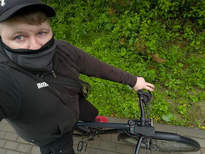 In an overhead shot, a person wearing a black baseball cap, a dark gray neck gaiter pulled up over their nose and mouth, and a black Puma hoodie is seated on a bicycle, holding the handlebars. The person's blue eyes are visible above the neck gaiter. The bicycle has a black frame and handlebars, with a display unit mounted on the handlebars. A black bag with a strap is worn across the person's chest. The background consists of lush green foliage and a paved walkway. The image is tagged with the location of Tarusa, Russia, suggesting the scene takes place within the city limits.
