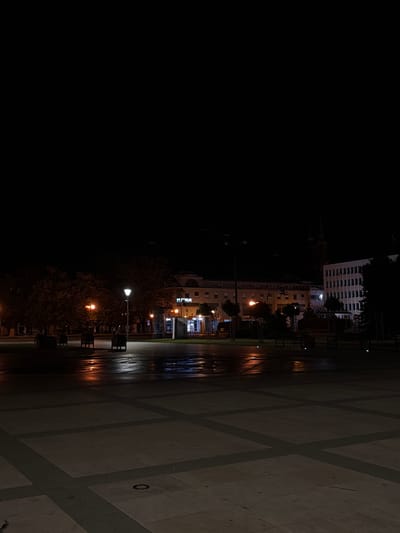 The image captures a dark, nighttime scene in the city of Siedlce, Poland. The foreground features a vast, paved plaza with a geometric pattern of large square tiles separated by darker grout lines. Several of these tiles are embedded with small, circular lights that emit a soft glow. In the mid-ground, a wet, reflective surface suggests recent rain, with warm-toned streetlights casting elongated reflections on the pavement. Scattered across this area are dark, rectangular planters or benches.
Further back, trees with dense foliage line the plaza. Beyond the trees, buildings are illuminated, indicating commercial or residential activity. One building in the mid-ground appears to be a multi-story structure with several windows and a prominent sign that reads "FITNESS" in bright blue lettering. To its right, another building shows a facade with many windows and a more imposing, modern architectural style. In the distance, the silhouette of a tall, spire-like structure, possibly a church tower, can be seen against the dark sky. The overall atmosphere is quiet and nocturnal, with the artificial lighting creating pools of illumination within the darkness.
