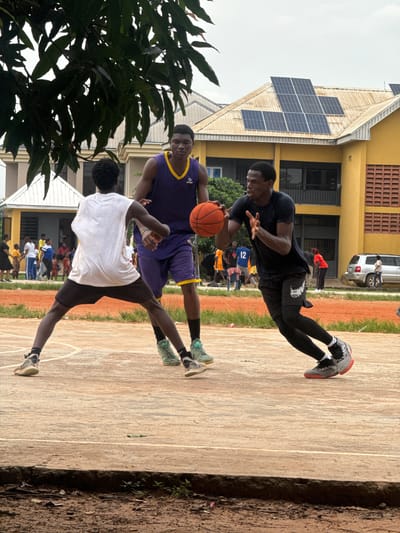 In Amansee, Nigeria, a basketball game is underway on an outdoor court. Three young men are the focus of the action. One man, wearing a purple basketball jersey and yellow trim, holds an orange basketball, preparing to pass or shoot. Facing him is another man in a white tank top and black shorts, his stance suggesting defense. A third man, dressed in a black t-shirt and black leggings, is dribbling the ball to his right, his body angled towards the basket. The court appears to be made of concrete with painted white lines. In the background, a yellow building with solar panels on its roof stands, and several other people, including children and adults, are visible watching the game or gathered nearby. A silver SUV is parked to the right of the building. The environment suggests a warm, possibly overcast day.
