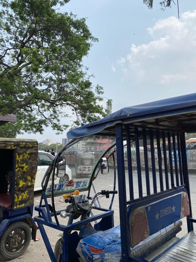 The image shows the view from inside an auto rickshaw in Narayanganj, Bangladesh. The foreground features the blue frame and parts of the rickshaw, including the steering mechanism, mirrors, and a camouflage-patterned side panel. Through the windshield, one can see other vehicles on a road, including cars and what appears to be another rickshaw. Buildings are visible in the background, indicating an urban setting. In the periphery, a tree with green leaves partially obscures the view on the left side, and the sky is visible above with some clouds. The overall impression is a snapshot of a typical street scene in Narayanganj.