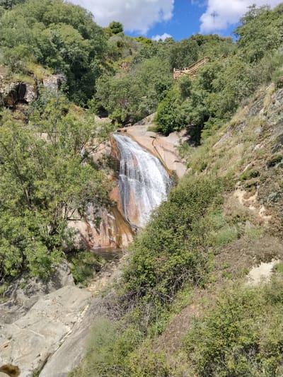 A medium shot depicts a waterfall cascading down a rock face surrounded by lush vegetation. The waterfall is the focal point, with water flowing over the reddish-brown stone. Green trees and bushes surround the falls, creating a natural enclosure. A wooden staircase or platform is visible in the upper right quadrant of the image, slightly obscured by the foliage. The sky is visible at the top, with blue patches and some clouds. The location is identified as Alijó, Portugal.