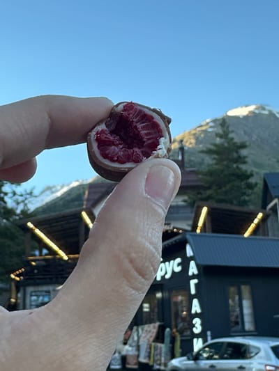 A close-up, slightly blurry photograph shows a person's thumb and index finger holding a chocolate-covered raspberry against a backdrop of mountains and buildings. The raspberry is halved, revealing its bright red interior. In the background, a dark-colored building with illuminated signs is visible, featuring Cyrillic lettering that appears to say "рус" and "МАГАЗИ". The building also has several windows and a dark roof. A silver car is parked in front of the building. Beyond the building, evergreen trees are scattered on the slopes of snow-capped mountains under a clear blue sky. The overall scene suggests a commercial area in a mountainous region, likely a resort town. The sign suggests the location might be a store or market in Terskol, Russia, given the Cyrillic script and mountainous setting.