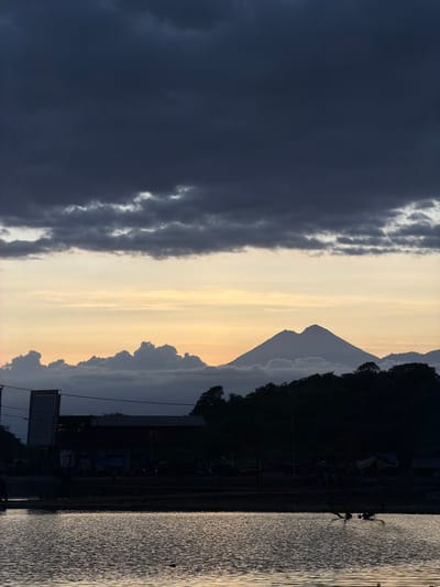 The image depicts a landscape at dusk or dawn, possibly located near Upt Tambak Sari, Indonesia. The sky is divided between a dark, stormy area on the left and a lighter, yellowish sky on the right. A silhouetted mountain range is visible in the center, with a distinct pointed peak. A dark, undefined area, potentially a body of water, occupies the right side of the image, reflecting the light. On the horizon, silhouetted buildings and structures are visible. The image is rotated 90 degrees counterclockwise.