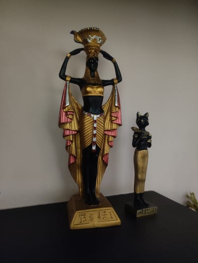 Two Egyptian-themed figurines are displayed on a dark, flat surface against a plain, light-colored wall. The larger figurine on the left depicts a standing woman with black skin, wearing a gold and red garment and carrying a gold headdress or vessel on her head. Her body is adorned with gold jewelry. The smaller figurine on the right represents a black cat-like figure, also standing and wearing a gold garment. Both figurines are detailed and appear to be made of a solid material. The overall scene is a still life arrangement. The image does not provide any information that would allow for a location in the city of Trikala, Greece.