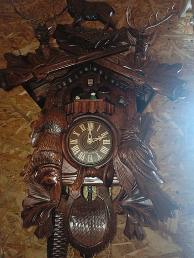 In Gorka, Russia, a large, ornate wooden cuckoo clock, featuring a dark brown finish and intricate carvings, is affixed to a light-colored, pressed wood panel wall. The clock's uppermost section displays a carved deer stag standing atop a platform of foliage. Below this, two carved deer heads with prominent antlers flank a pair of crossed hunting rifles or horns. Directly beneath the crossed implements, a small balcony houses two small, articulated figures: one predominantly white with dark spots, and another with a green body and black hat, positioned next to a small red object. Below the balcony, the central circular clock face features white Roman numerals, gold-toned hands, and the inscription "MADE IN GERMANY." The time displayed is approximately 1:55. Flanking the clock face are large, deeply carved predatory birds, with additional foliage and smaller bird carvings on the left side. A carved hunting horn is positioned horizontally beneath the clock face. The lower section of the clock is adorned with a large, textured carved shield or pouch, with a carved pine cone weight suspended by a chain visible below it. Another chain is partially visible to the right of the shield. The overall scene is illuminated with a strong light source originating from the right, causing a bright glare.